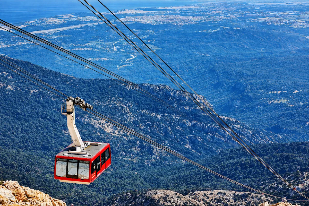 Kırmızı bir teleferik kabini, Olympos Teleferik hattında Tahtalı Dağı'nın yeşil ormanlarla kaplı yamaçları üzerinden süzülürken, uzaklarda Antalya'nın masmavi denizi ve kıyı şeridinin muhteşem panoramik manzarasını sunuyor.