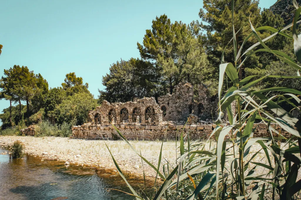 Antalya gezi: Olympos Antik Kenti'nin ağaçlarla çevrili kıyısında, kemerli pencereleri belirgin taş duvar kalıntıları, yemyeşil doğanın içinde berrak bir akarsuyun kenarında yer alarak Antalya'nın tarihi ve doğal güzelliklerini bir araya getiren büyüleyici bir manzara sunuyor.