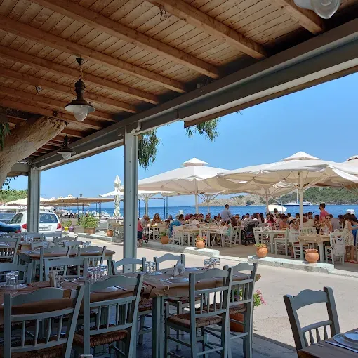 Güneşli bir günde, ahşap tavanlı açık hava restoranının ön planında boş masalar ve açık mavi sandalyeler görülürken, sağ tarafta beyaz şemsiyeler altında masmavi denize nazır yemek yiyen kalabalık aileler ve insanlar, arka planda ise sakin bir koy ve tekne manzarasıyla keyifli bir sahil restoranı ortamı.