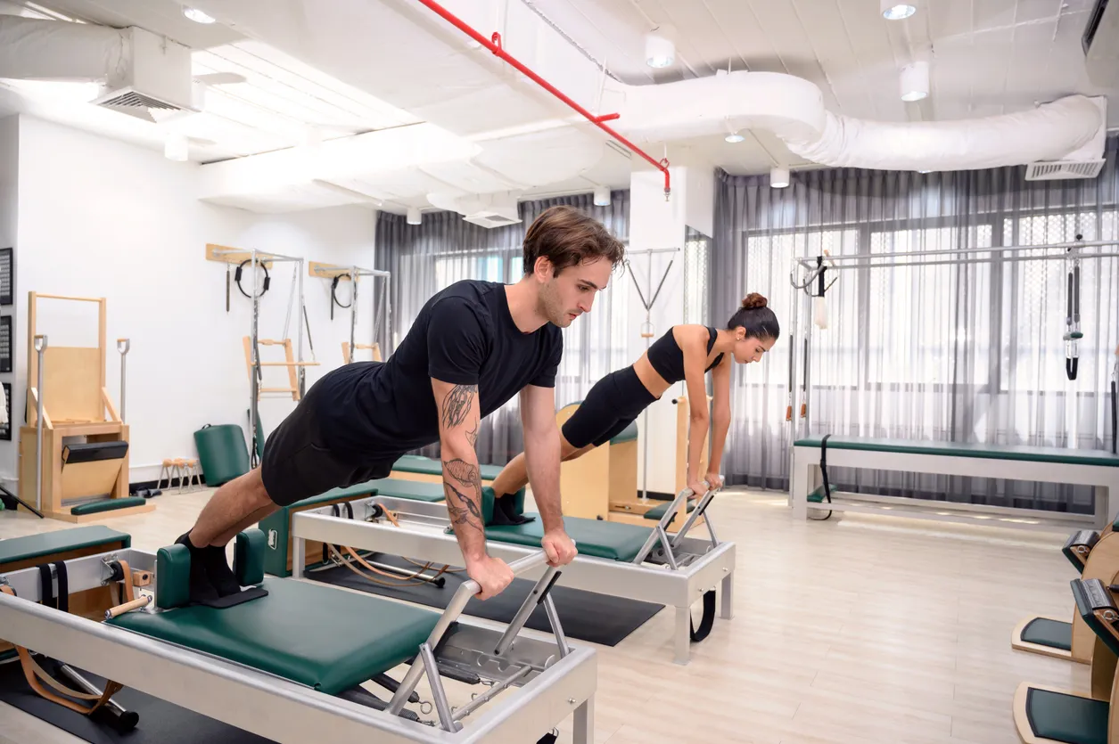 Aydınlık bir pilates stüdyosunda, genç bir adam ve kadın, başlangıç seviyesi pilates dersine uygun olarak reformer aletleri üzerinde kol ve karın kaslarını çalıştıran bir plank pozisyonunda dengede durarak egzersiz yapıyorlar.