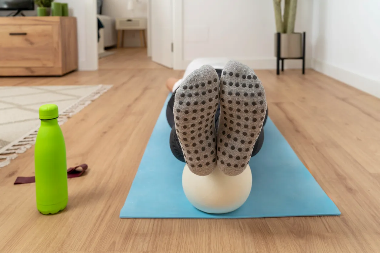 Mavi yoga matı üzerinde dinlenen bir kişinin, tabanlarında siyah kaydırmaz noktalar bulunan gri pilates çorapları giymiş ayakları, egzersiz sırasında kullanılan küçük, açık renkli bir topun üzerinde rahatça duruyor; yan tarafta parlak yeşil bir su matara dikkat çekiyor.