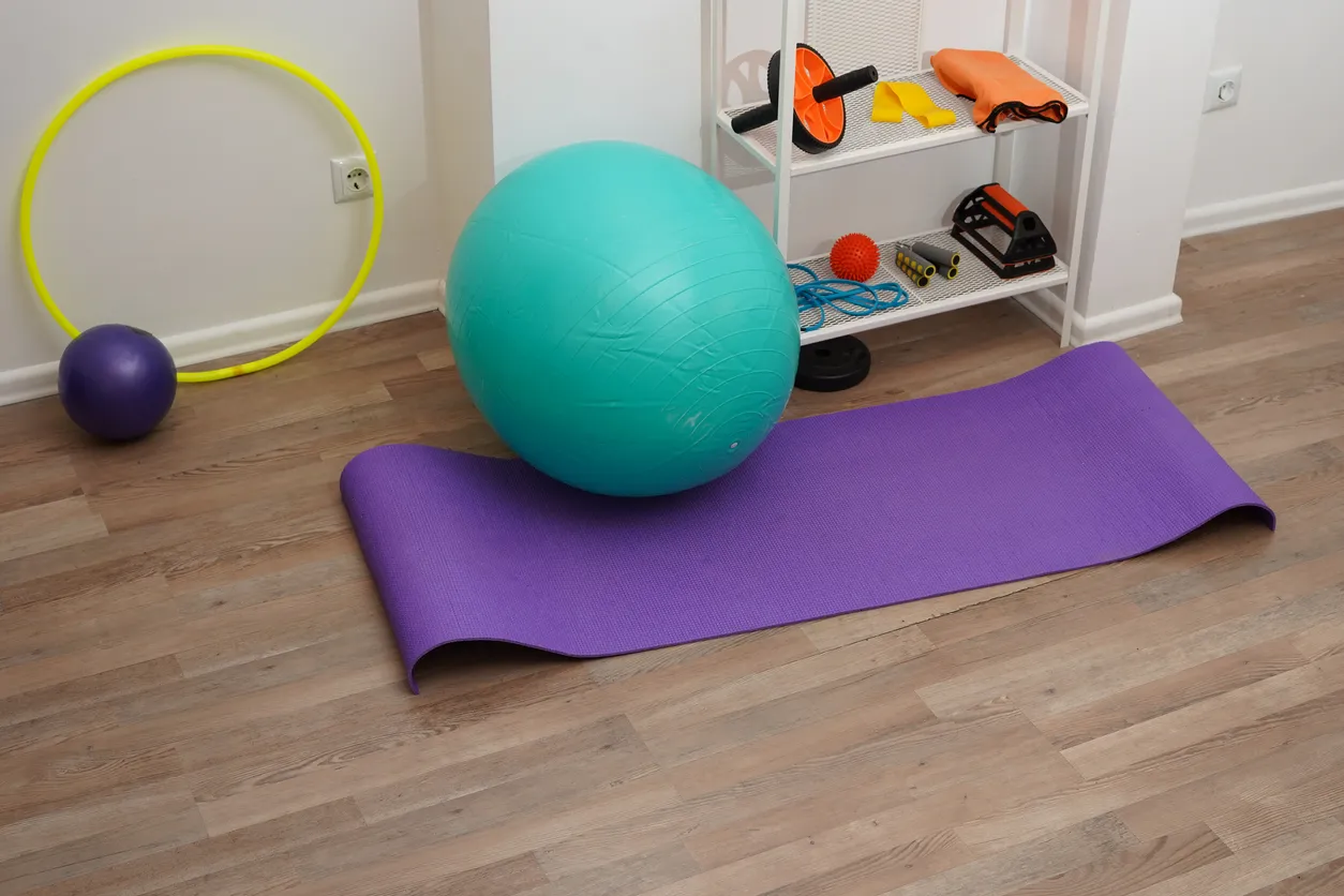 Ahşap zemin üzerinde, mor bir matın üzerinde duran büyük turkuaz bir pilates topu, sarı bir hulahop, küçük mor bir top ve beyaz bir rafta duran çeşitli diğer pilates ekipmanları (mekik aleti, atlama ipi, direnç bandı) beyaz duvar önünde düzenlenmiş bir şekilde görünüyor.