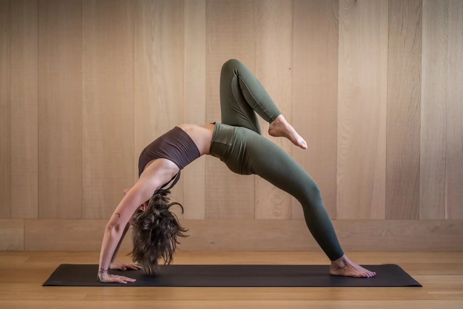 Kadın bir pilates matının üzerinde, koyu kahverengi spor sütyeni ve haki yeşili taytıyla esnekliğini sergileyen zorlu bir pilates egzersizi yapıyor; sırtını bir köprü gibi kavisli hale getirip sağ bacağını dizden bükerek yukarı kaldırmış, bu pozisyon pilateste nasıl daha güçlü ve esnek olunacağını gösteriyor.