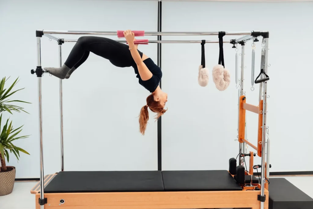 Cadillac pilates aletinde baş aşağı ters bir pozisyonda, siyah taytlı ve siyah tişörtlü bir kadın, elleriyle barı tutarken bacaklarını bükerek dengeli ve gelişmiş bir pilates egzersizi sergiliyor.