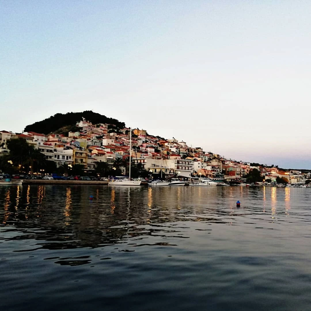 Alacakaranlıkta Plomari'nin Midilli Adası sahilinde, yamaç boyunca sıralanmış kırmızı çatılı ve pastel renkli binaların ışıklarının sakin denize yansırken demirli teknelerle bütünleştiği huzurlu bir görünüm.