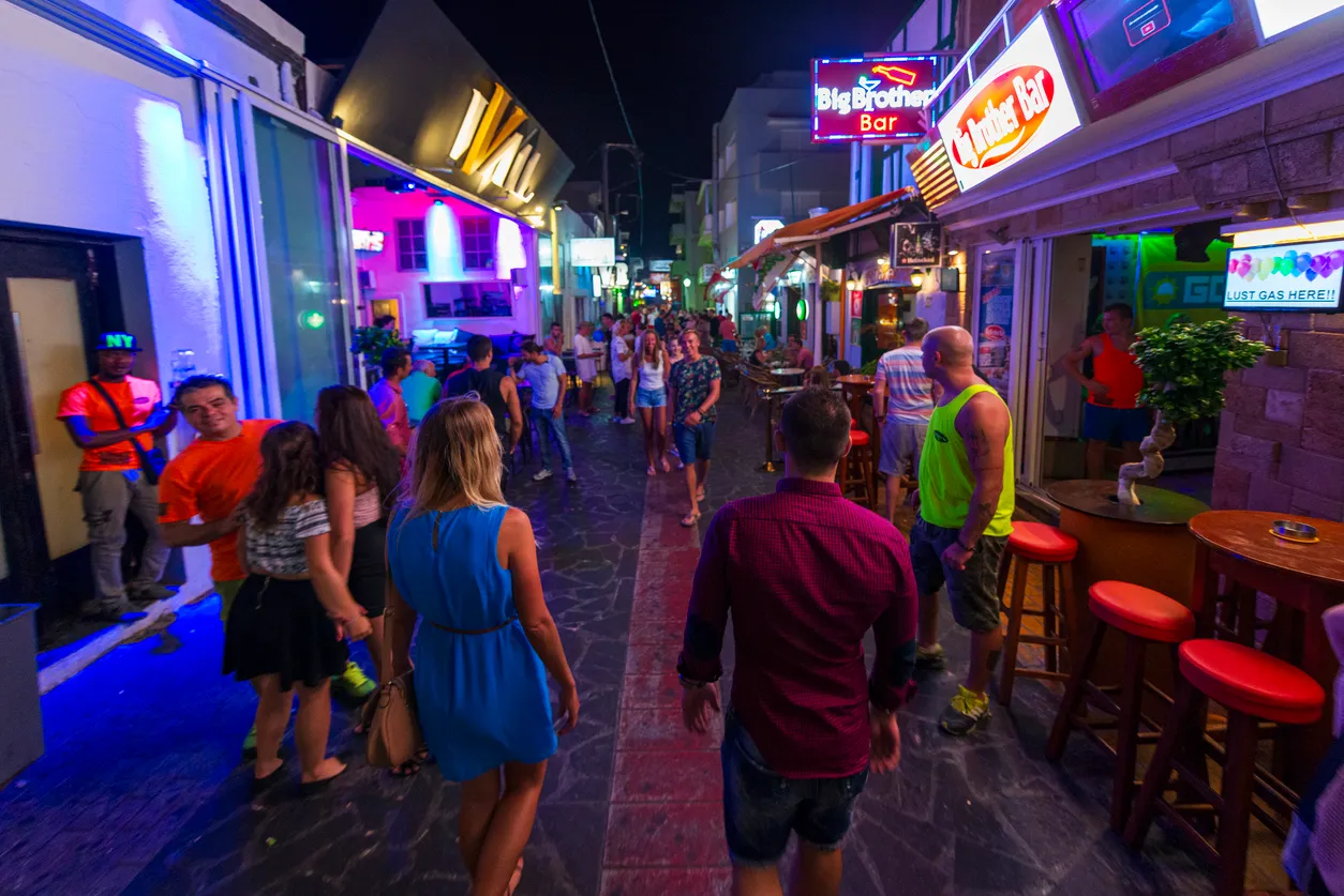 Rodos'un canlı gece hayatını yansıtan bu geniş açılı görselde, cadde boyunca uzanan renkli neon tabelalı barların ve eğlence mekanlarının önünde yürüyen, sohbet eden ve keyifli vakit geçiren çeşitli yaş gruplarından kalabalık insanlar görülüyor.