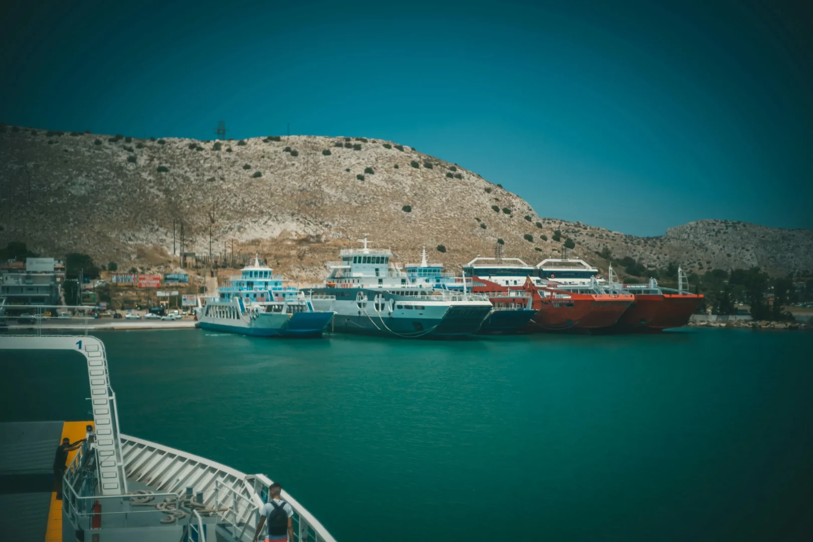 Seyir halindeki bir feribotun güvertesinden çekilen bu görselde, berrak turkuaz deniz üzerinde, arka planda uzanan dağlık kıyı şeridi önünde sıralanmış, farklı renk ve boyutlardaki çok sayıda feribotun yanaştığı canlı bir liman manzarası görülmekte, bu da Rodos'a feribot ile ulaşım için önemli bir geçiş noktasını vurgulamaktadır.