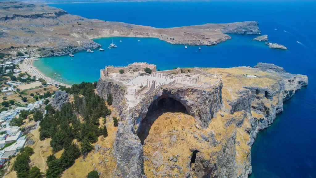 Rodosa yolculuk edenler için muhteşem bir manzara sunan, kayalık bir burun üzerinde Lindos Akropolisi'nin antik kalıntıları, altında turkuaz renkli bir koyda demirlemiş tekneler ve şemsiyelerle dolu kumlu plajı ile kuşbakışı görünüyor.