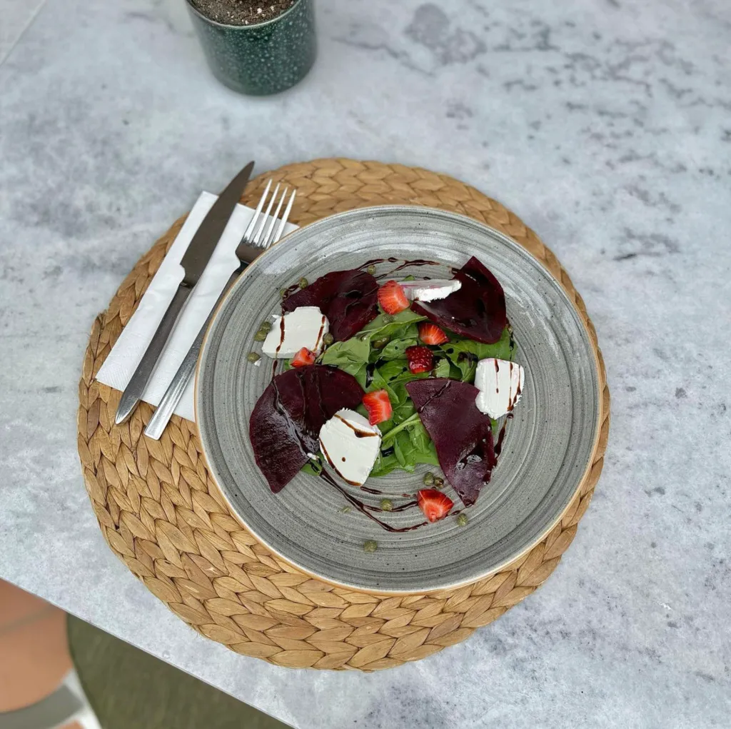 Hasır servis üzerinde gri tabakta sunulmuş, roka, çilek, keçi peyniri ve ince dilimlenmiş pancarla hazırlanmış taze salata.