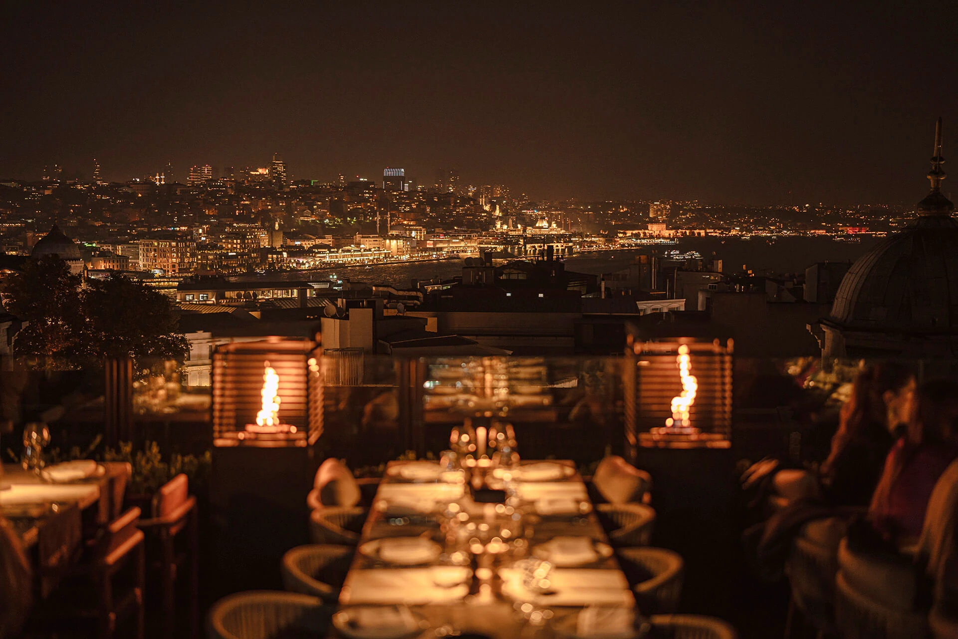 Gece, İstanbul'un parıldayan panoramik şehir manzarasına nazır bir teras restoranında, sıcak bir ambiyans yaratan alevli dış mekan ısıtıcılarının ışığında, özenle hazırlanmış masaların şık akşam yemeği atmosferi.