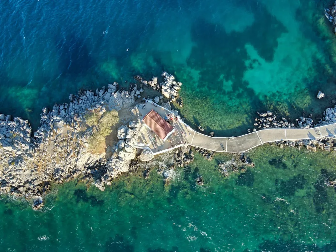 Sakız Adası'nda, berrak turkuaz suların ortasında, kayalık bir adacık üzerinde kırmızı çatılı küçük bir yapının bulunduğu, kıvrımlı bir iskele ile karaya bağlanan pitoresk bir koyun kuşbakışı görüntüsü.
