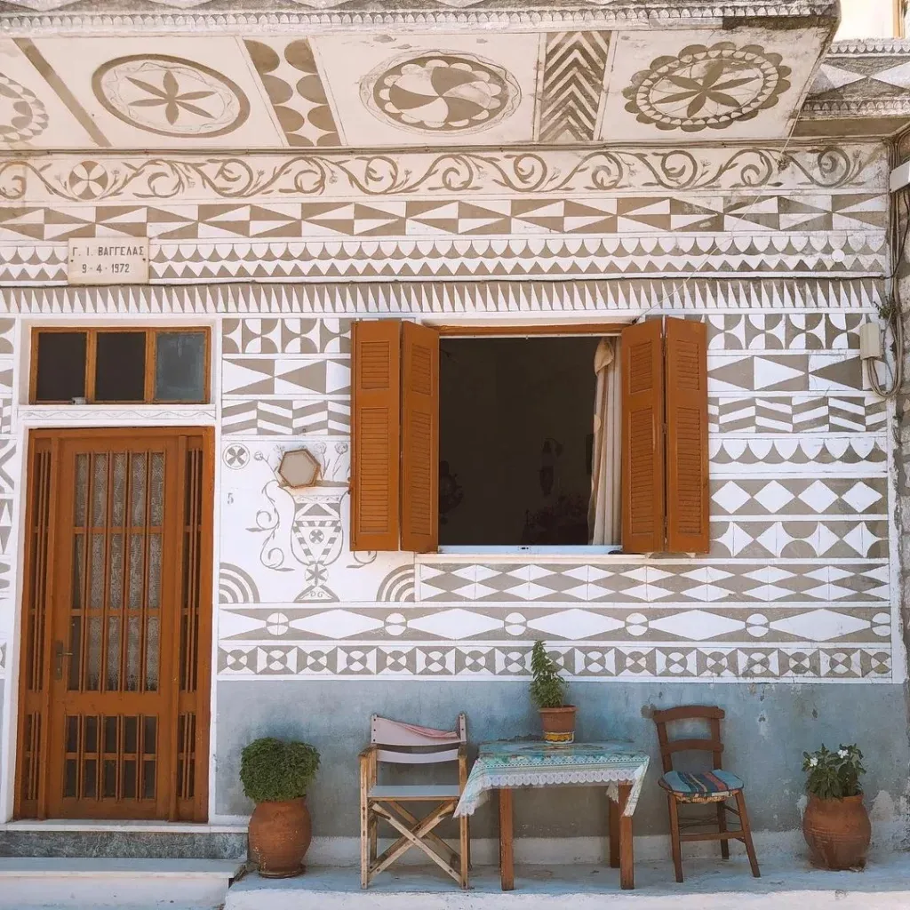 Sakız Adası'nda gezilecek yerlerden biri olan bu binanın dış cephesi, kahverengi ahşap kapısı ve pencereleriyle gri ve beyaz geometrik desenlerle süslenmiş olup, önündeki küçük masa ve sandalyelerle dinlendirici bir oturma alanı sunuyor.