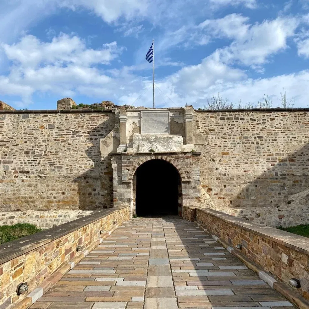 Sakız Adası'nın tarihi dokusunu yansıtan bir gezilecek yer olan taş bir kalenin kemerli ana girişine, üzerinde dalgalanan Yunan bayrağı ve gökyüzündeki beyaz bulutlarla birlikte uzanan geniş, taş döşeli bir köprü görülüyor.