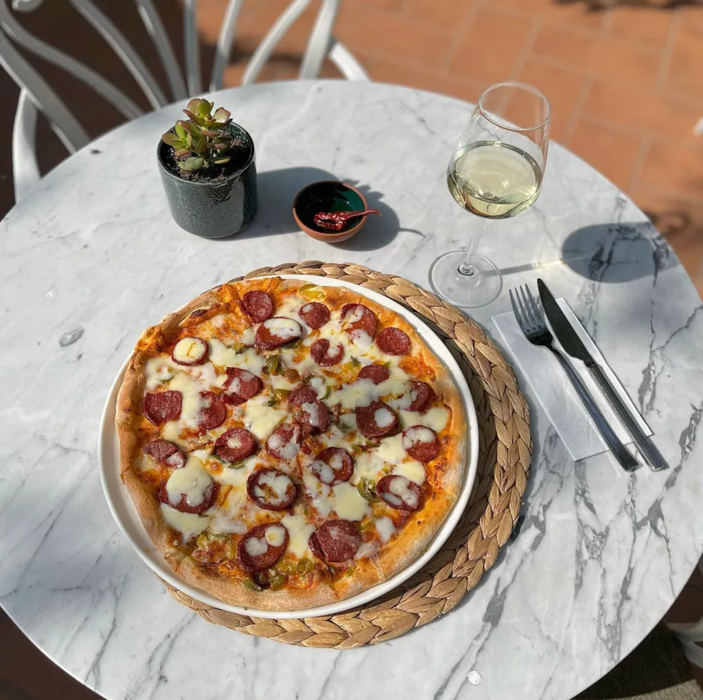 Mermer masa üzerinde servis edilen sucuklu pizza, yanında beyaz şarap kadehi ve çatal bıçak takımıyla güneşli bir ortamda sunulmuş.