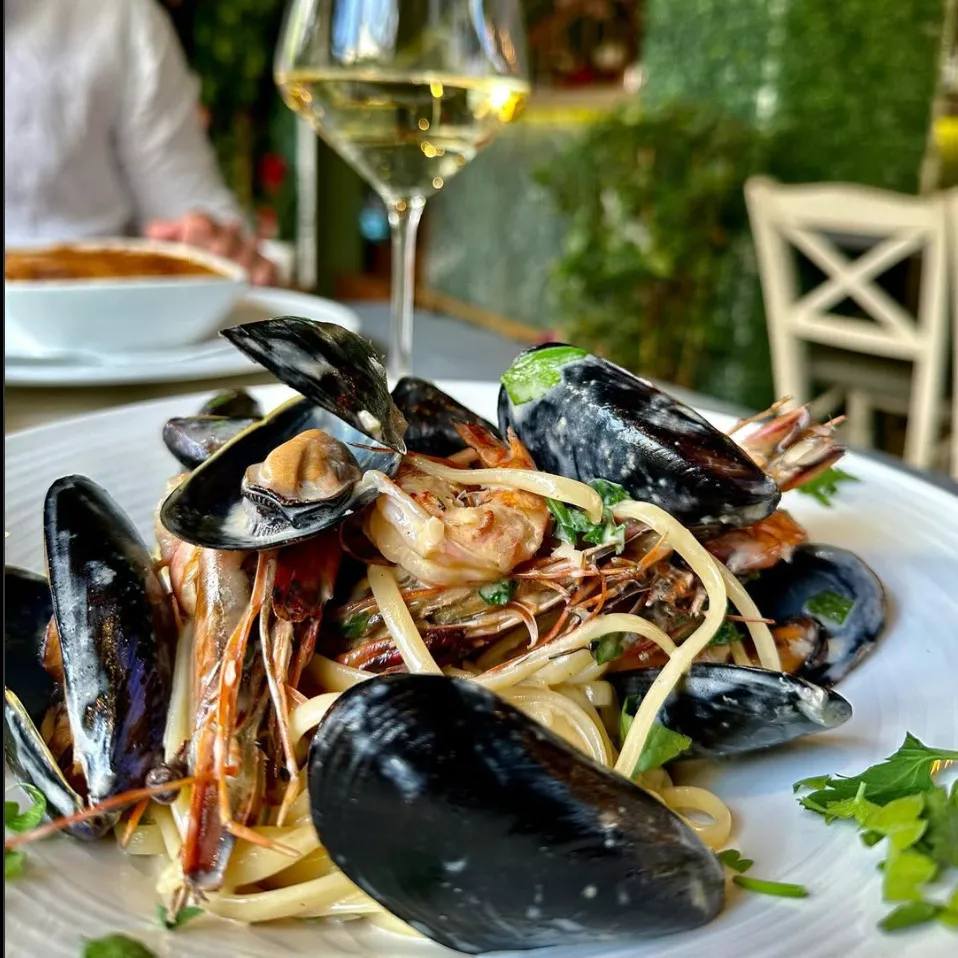 Bu yakın çekim fotoğraf, Rodos'taki Tamam Restaurant'ın nefis deniz mahsullü makarnasını, kabuklu midyeler, karidesler ve linguine ile birlikte, arka planda bir kadeh beyaz şarap ve bulanık bir restoran ortamıyla gösteriyor.