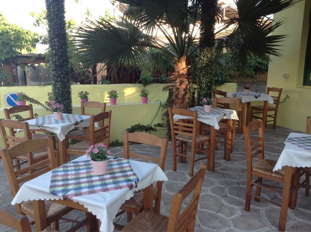 Açık havada, taş zeminli bir taverna avlusunda, beyaz ve ekose desenli masa örtüleri serilmiş ahşap masalar ile hasır sandalyeler bulunuyor; her masada pembe saksı çiçekleri, arka planda palmiye ağaçları ve sarı duvar hoş bir atmosfer yaratıyor.