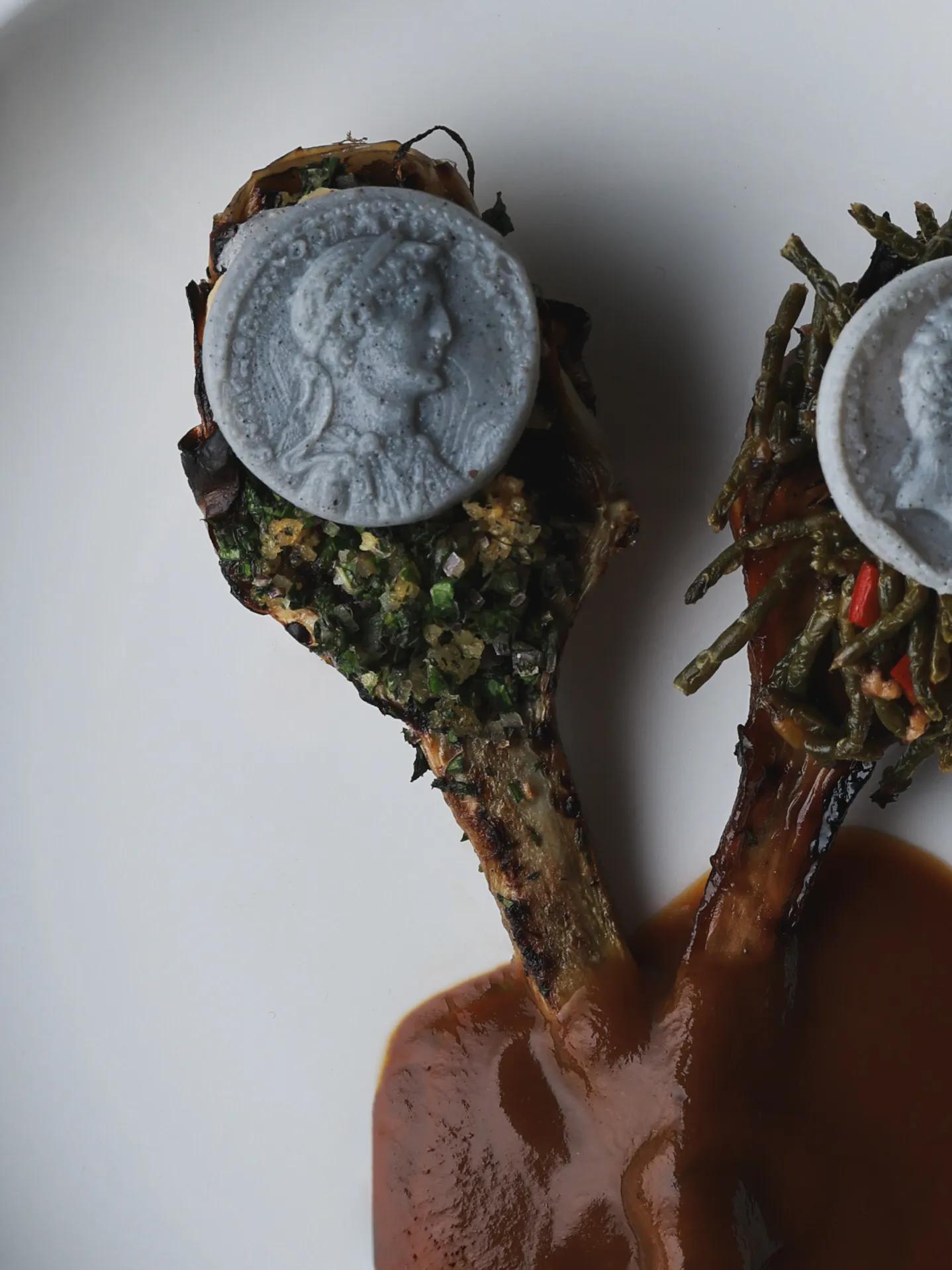Telezzüz'ün doğal malzemelerle hazırlanan vegan fine dining konseptini vurgulayan bu görselde, üzerinde antik motifli gri yenilebilir madalyonlar bulunan, birinde yeşil otlar, diğerinde ise uzun yeşil sebzelerle zenginleştirilmiş iki adet ana yemek, zengin kahverengi bir sos eşliğinde beyaz tabakta zarifçe sunuluyor.