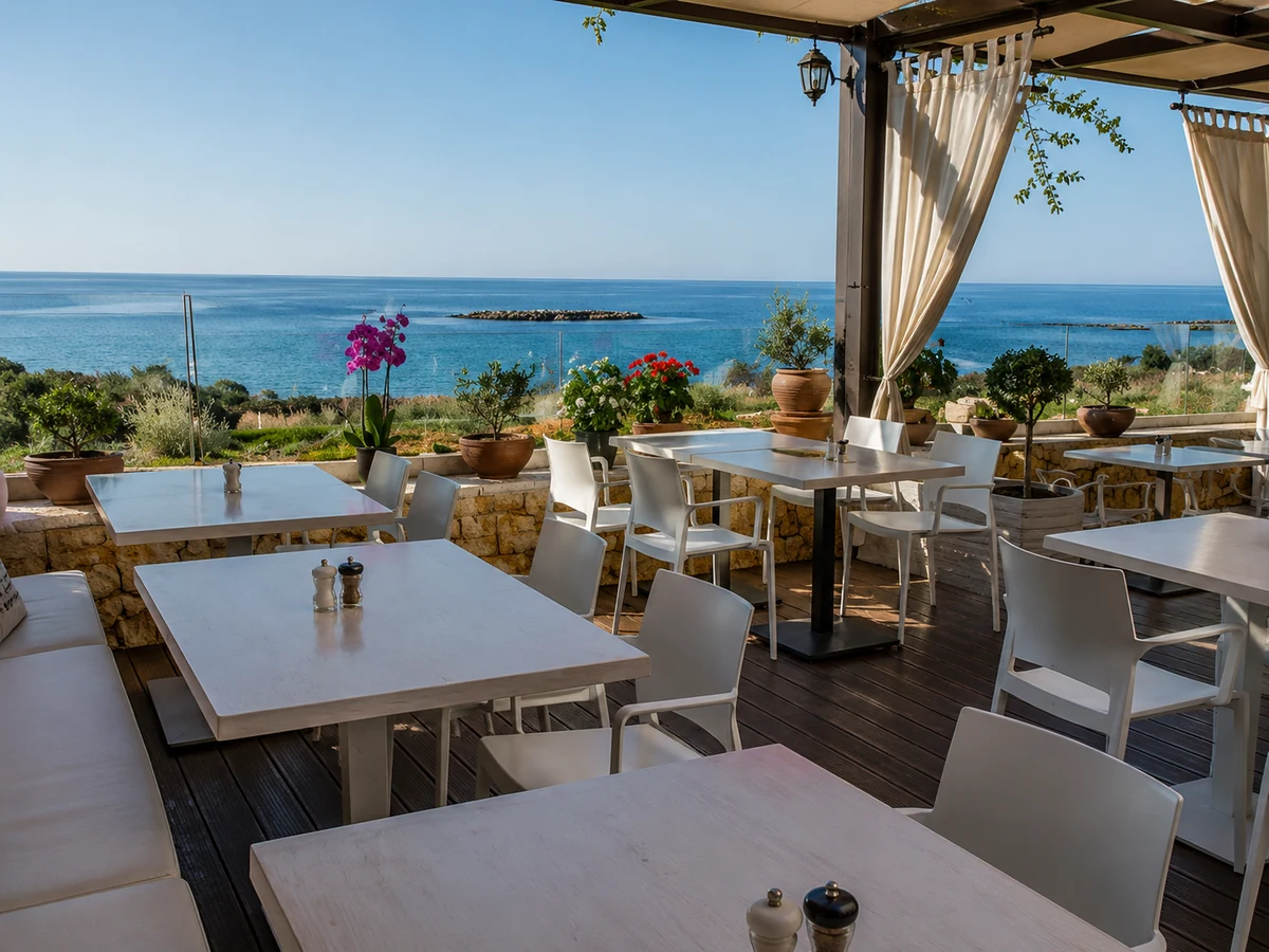 Theodosi Girit Restoranı'nın Ege Denizi'ne bakan, beyaz masalar, sandalyeler ve mor orkide gibi renkli bitkilerle donatılmış, huzurlu ve geniş açık hava terası, misafirlerine ferah bir deniz manzarası eşliğinde keyifli bir yemek deneyimi sunuyor.