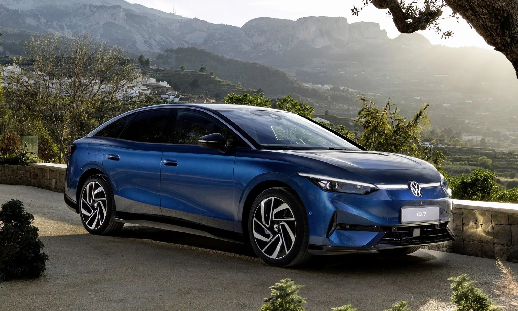Koyu mavi renkli, şık bir Volkswagen ID.7 elektrikli otomobil, dağlık bir manzara, yeşil bitki örtüsü ve beyaz evlerin görüldüğü kırsal bir yerleşim yerinin önünde, güneşli bir havada hafifçe sağa dönük bir açıyla park edilmiş, modern tasarımıyla adeta yeni nesil bir elektrikli Passat izlenimi veriyor.