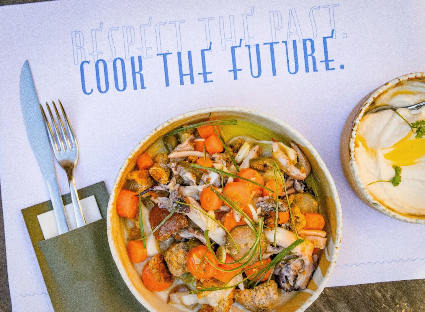 Üzerinde RESPECT THE PAST. COOK THE FUTURE. yazan beyaz bir Amerikan servis üzerinde, karides, kalamar, midye, kızarmış ekmek parçaları, küp havuç, turşu dilimleri ve taze otlarla zenginleştirilmiş, geçmişin ve geleceğin lezzetlerini harmanlayan modern bir deniz ürünleri yemeği, yanında zeytinyağlı kremalı bir meze kasesi ve şık çatal bıçak takımıyla iştah açıcı bir şekilde sunuluyor.