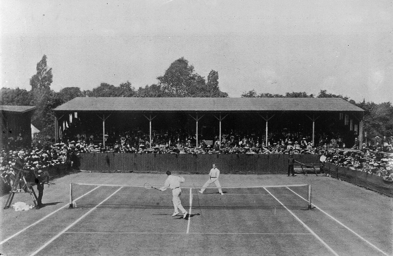 Wimbledon'ın 1910 yılındaki bir karşılaşmasını gösteren bu tarihi siyah beyaz fotoğrafta, beyaz tenis kıyafetleri içindeki iki sporcu çim kortta raket sallarken, arkalarındaki ahşap çatılı tribünü dolduran şapkalı ve şık giyimli seyirciler oyunu dikkatle izliyor.
