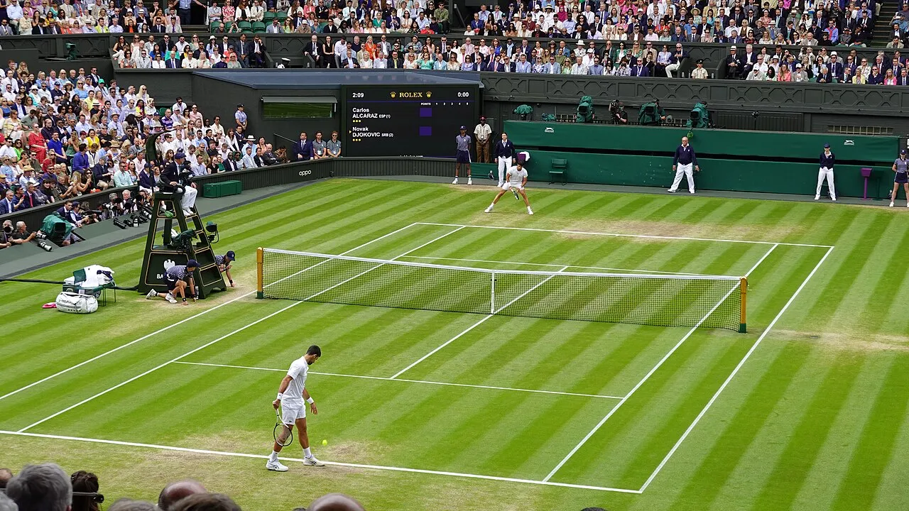 2023 Wimbledon tenis maçında, çim kortun ön planında raketiyle topa bakan Novak Djokovic, file arkasında ise hazır bekleyen Carlos Alcaraz, kalabalık seyirci tribünlerinin çevrelediği şeritle çim sahada mücadele ediyorlar.