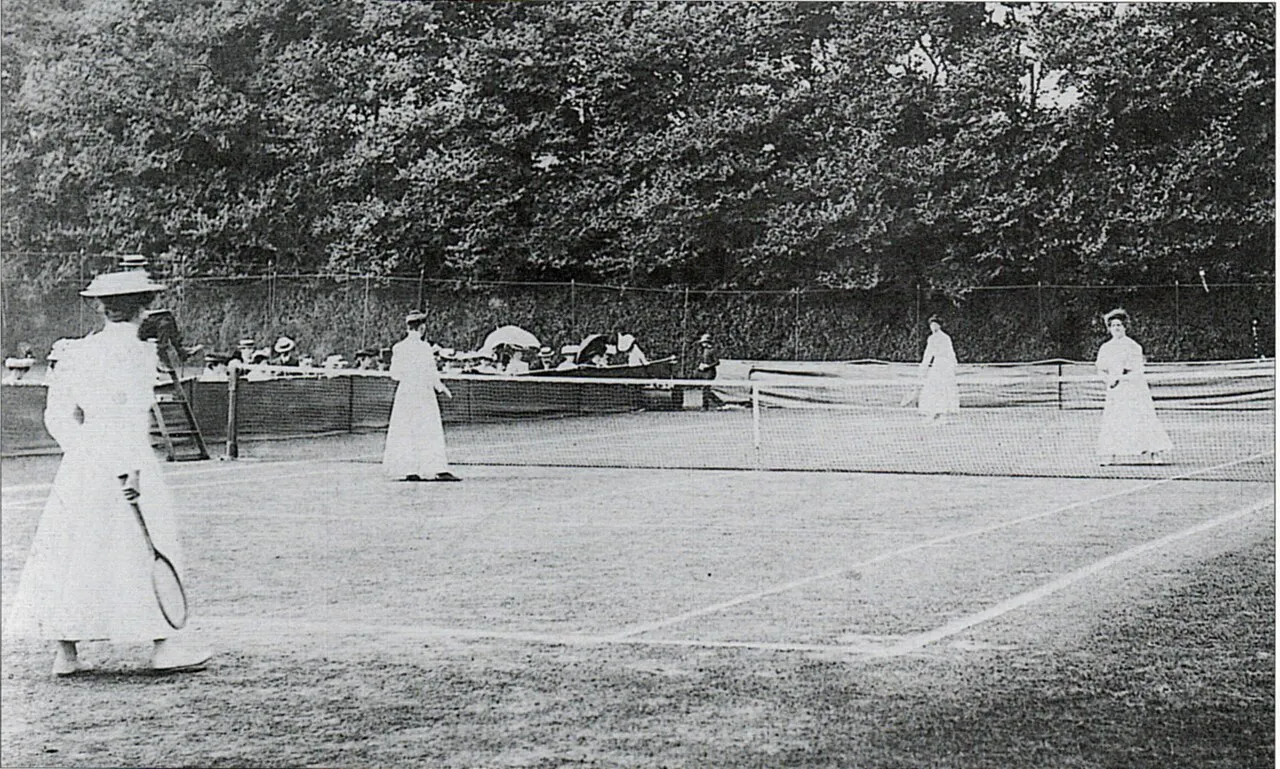 Wimbledon'ın erken dönem tarihinden siyah beyaz bir fotoğraf, uzun beyaz elbiseleri ve şapkalarıyla çim kortta raketleriyle zarifçe tenis oynayan kadınları ve arka plandaki seyircileri gösteriyor.