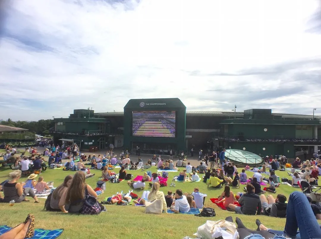 Wimbledon'daki ünlü Henman Tepesi'nde, bulutlu bir gökyüzünün altında, yeşil çimenlerin üzerine serilmiş battaniyelerin üzerinde uzanmış ve oturmuş yüzlerce kişi, The Championships turnuvasının tenis maçını dev bir ekrandan keyifle izleyerek açık hava atmosferinin tadını çıkarıyor.