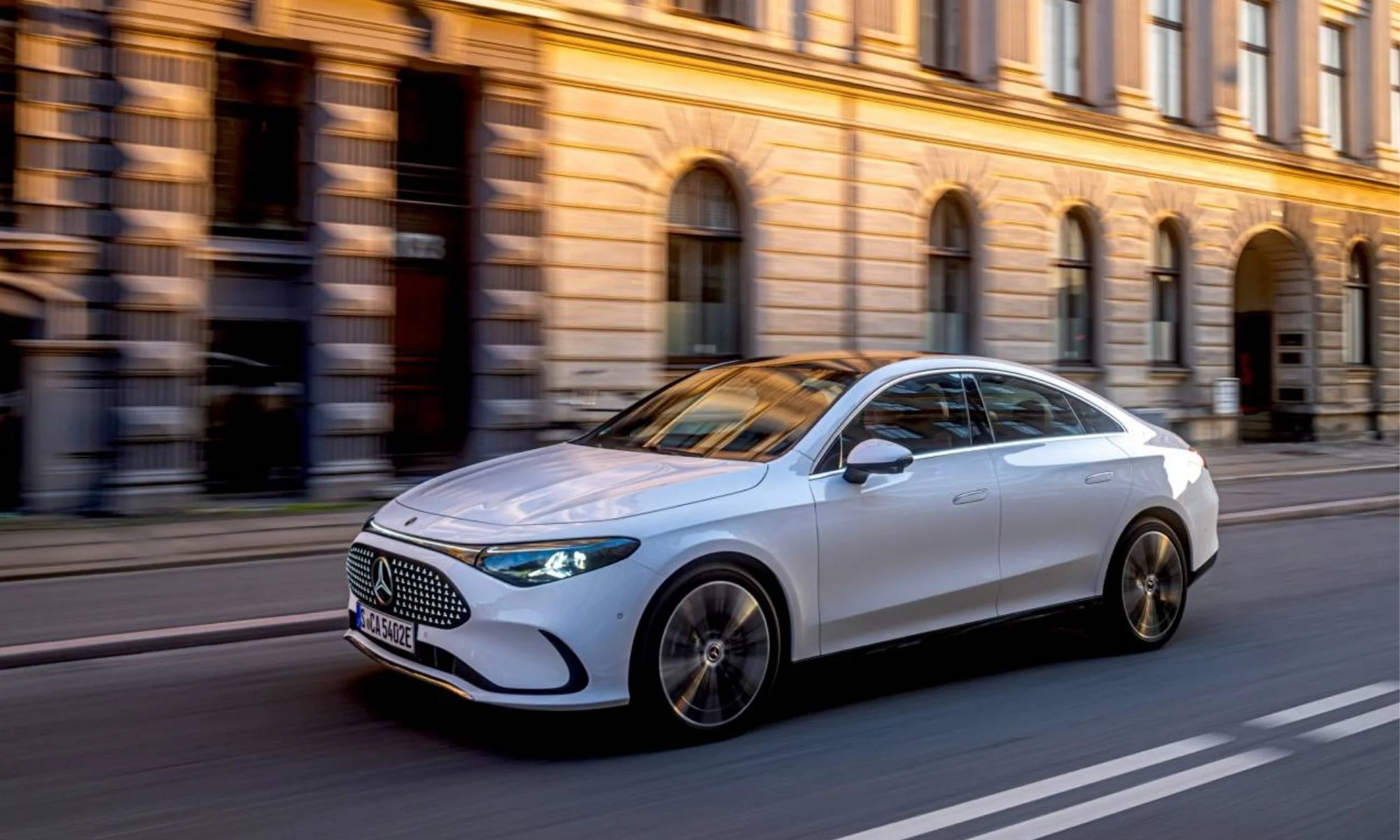 Güneşli bir şehir caddesinde ilerleyen, beyaz renkli, dinamik hatlara sahip, modern ön ızgarası ve parlak LED farlarıyla dikkat çeken bir Mercedes-Benz elektrikli sedan otomobil, arka plandaki hareket bulanıklığıyla hız hissi veriyor.