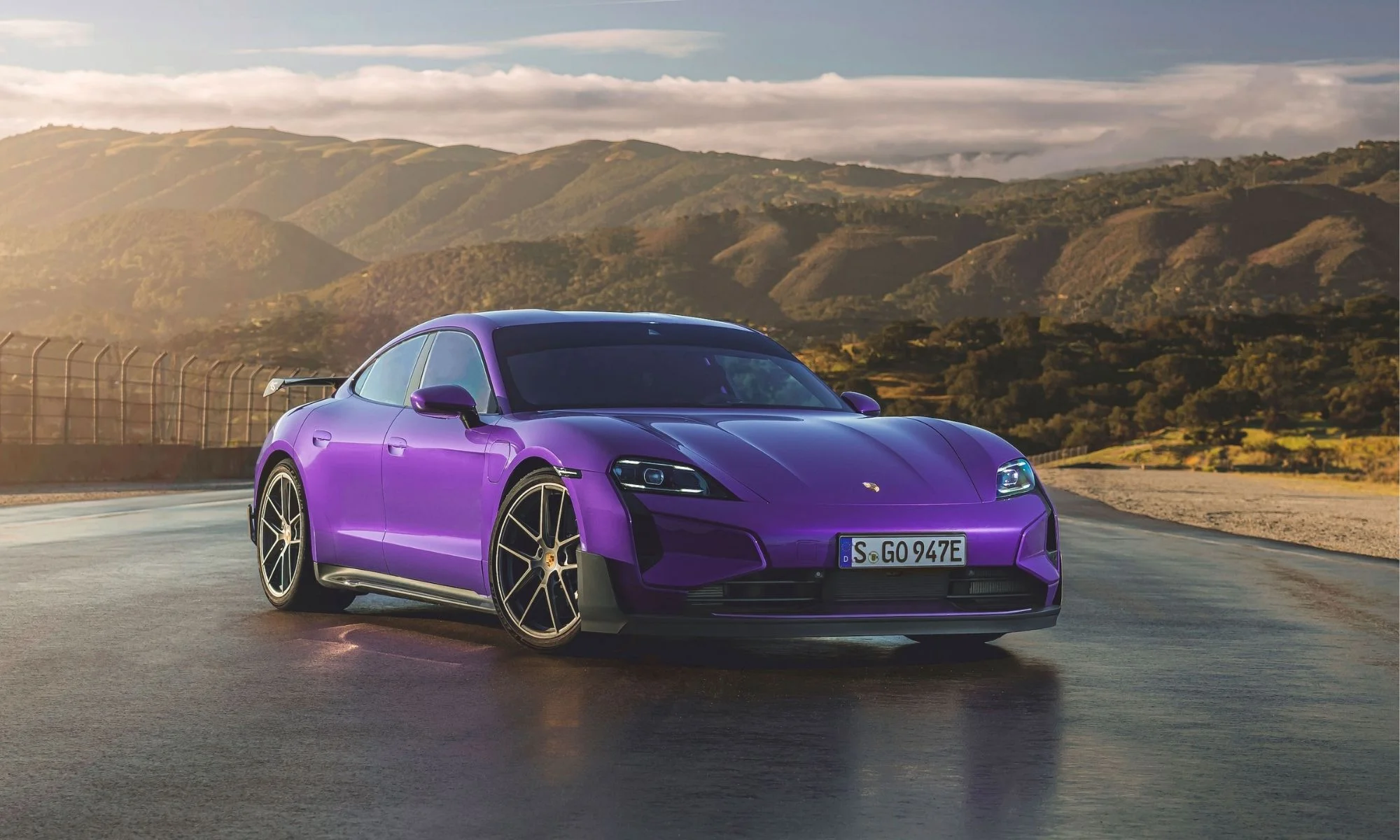 Ön sağ çaprazdan çekilmiş bu fotoğraf, ıslak bir asfalt yol üzerinde duran mor renkli, makyajlı yeni Porsche Taycan'ı gösteriyor; arka planda güneş ışığıyla aydınlanmış tepeler ve bulutlu bir gökyüzü, otomobilin yenilenen tasarımını ön plana çıkarıyor.