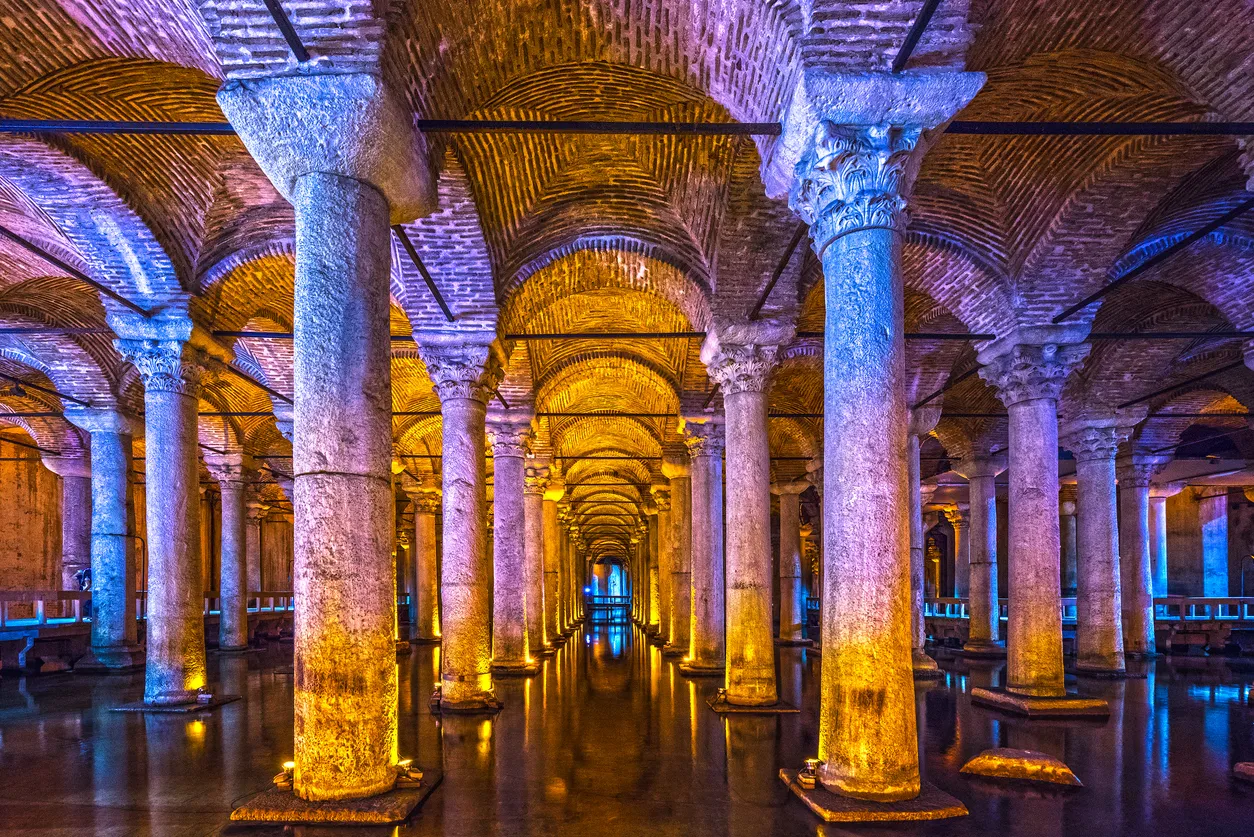 Yerebatan Sarnıcı'nın restorasyon sonrası göz alıcı atmosferinde, suyla kaplı zeminde yansıyan mor, mavi ve altın tonlarındaki ışıklarla aydınlatılmış, sıralı sütunların ve kemerli tavanın oluşturduğu uzun ve büyüleyici bir geçit manzarası.