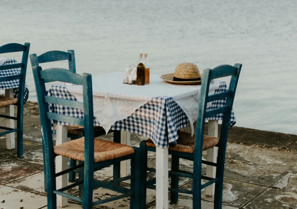 Zakintos gezilecek yerler arasında yer alan huzurlu bir taverna sahnesi; deniz kenarında, mavi tahta sandalyelerin ve hasır şapkanın eşlik ettiği beyaz örtülü masada zeytinyağı şişeleriyle keyifli bir mola imkanı sunuyor.