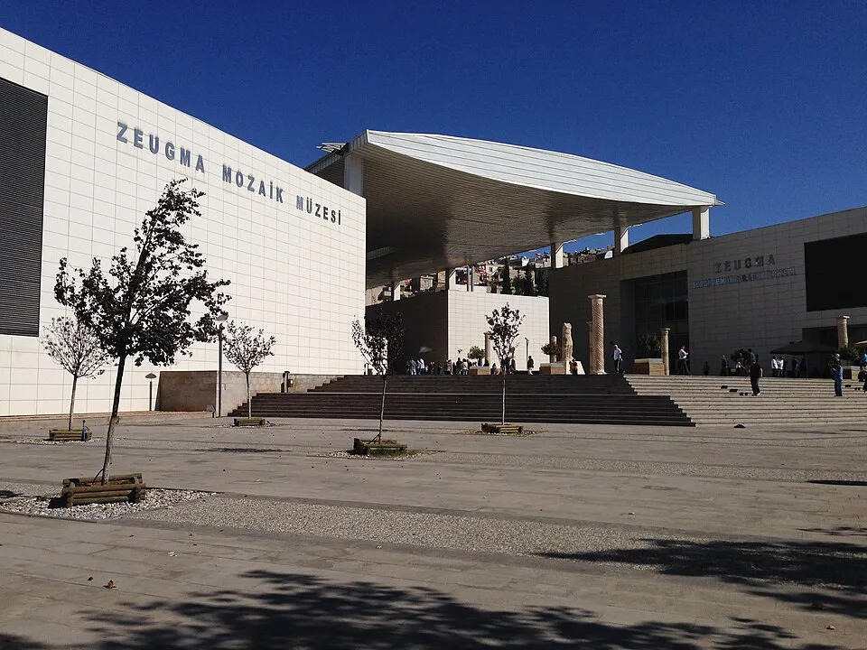 Güneşli bir havada, ZEUGMA MOZAİK MÜZESİ tabelasıyla dikkat çeken modern binanın önündeki geniş meydanda, beyaz fayanslı binaya çıkan merdivenlerde ziyaretçiler ve binanın altında antik sütun benzeri yapılar ile büyük bir gölgelik görülebiliyor.