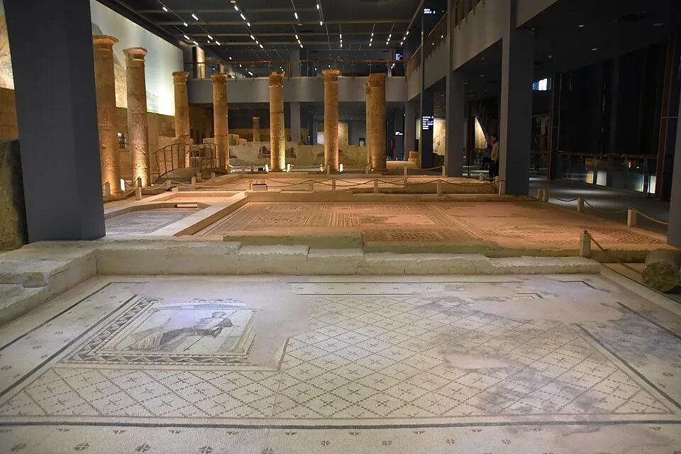 Zeugma Mozaik Müzesi'nin etkileyici geniş bir salonunda, yerden ışıklarla aydınlatılan yüksek antik sütunlar arasında, ön planda insan figürlü detaylı bir mozaiğin ve arka planda farklı geometrik desenlere sahip çok sayıda korunmuş mozaik zeminin sergilendiği bir atmosfer betimleniyor.
