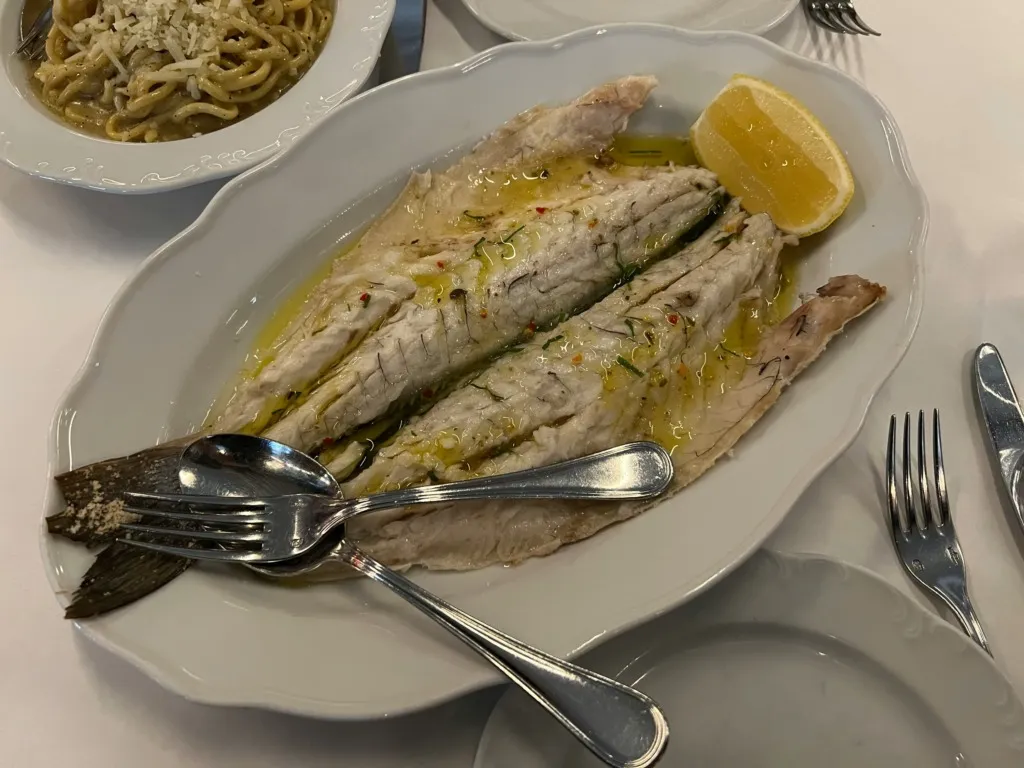 Zeytinyağlı sos ve limon eşliğinde servis edilmiş beyaz etli balık filetosu, yanında rendelenmiş peynirli makarna ile bir restoranda sunuluyor