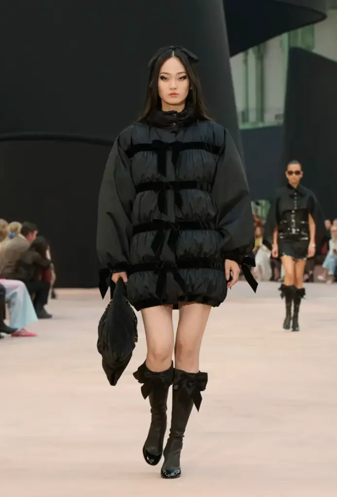 Siyah volan detaylı puffer mini elbise, kurdeleli çizmeler ve yumuşak hatlı aksesuarlarla kombinlenerek modern gotik bir couture stili yaratıyor.