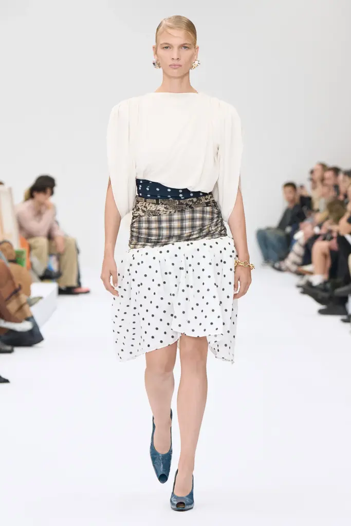 Acne Studios'un ilkbahar/yaz 2025 defilesinden bir görünümde, podyumda yürüyen bir manken, hacimli, krem rengi bir üst, lacivert puantiyeli ve ekose kumaşlardan oluşan katmanlı bir bel detayı ile siyah puantiyeli asimetrik beyaz bir etek ve parlak lacivert açık burunlu topuklu ayakkabılar giyiyor.