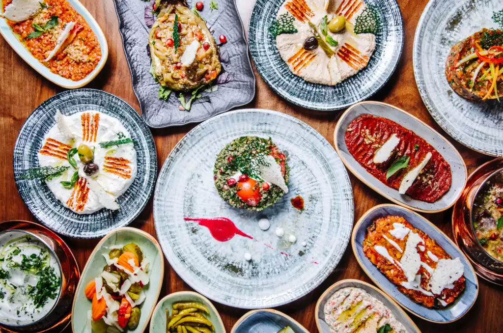 Ahşap bir masa üzerinde, humus, haydari, patlıcan salatası ve nar taneleriyle süslenmiş yeşillik gibi birçok farklı ve rengarenk meze tabağı, Akyaka ve çevresindeki kıyı bölgelerinde deneyimlenebilecek taptaze lezzetlerle dolu zengin bir Ege sofrasını gözler önüne seriyor.