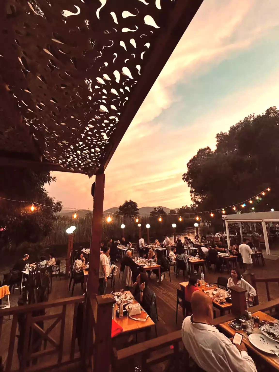 Gün batımının turuncu ve pembe tonlarıyla aydınlanan gökyüzünün altında, Akyaka'daki bir restoranın ahşap iskelesinde toplanmış, asılı ışıkların ve üst kısımdaki dekoratif tavanın oluşturduğu sıcak ambiyansta yemek yiyen kalabalık bir misafir topluluğu.