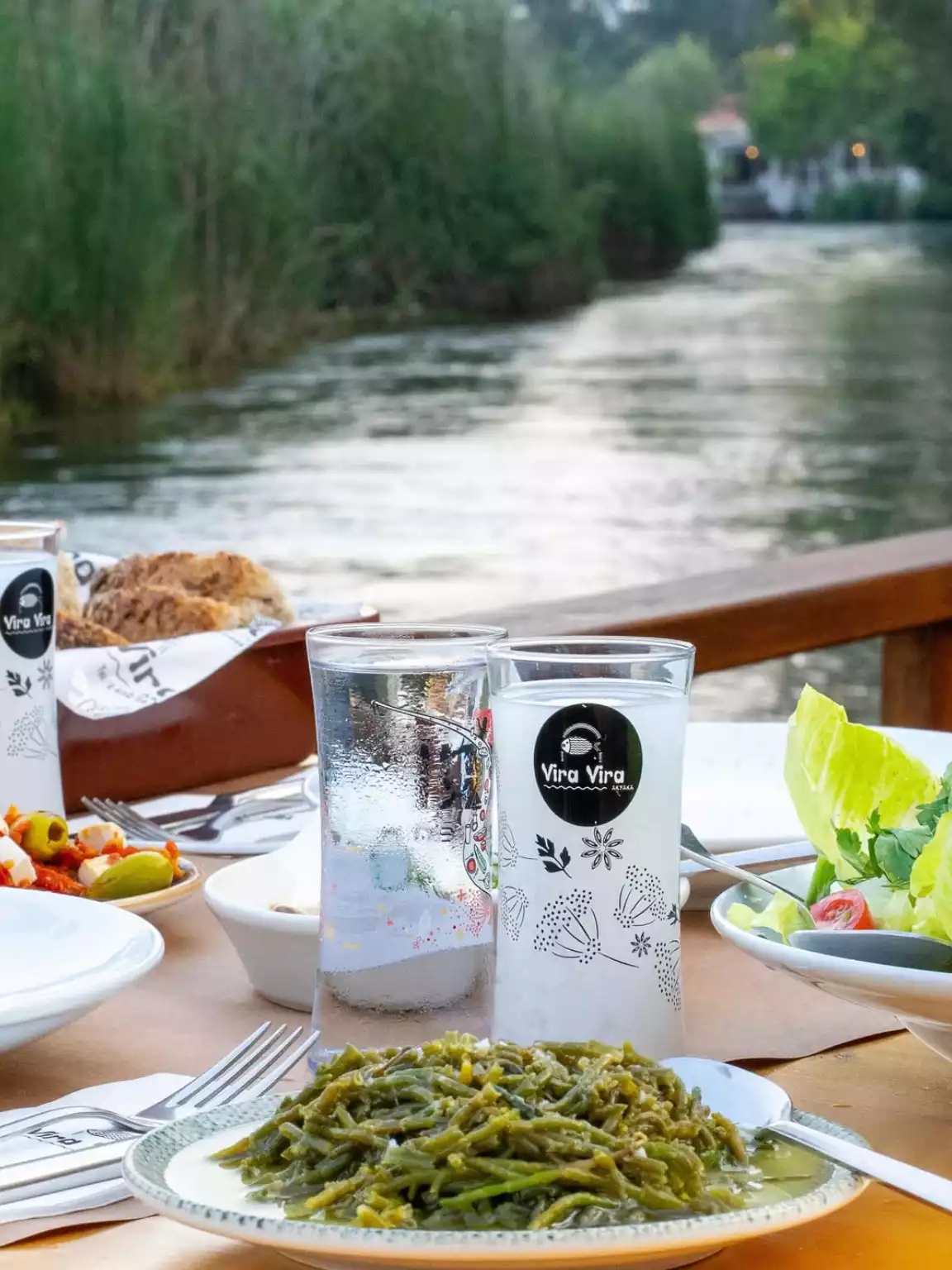 Akyaka'nın berrak sularıyla ünlü Azmak Nehri kenarında, yeşil bitki örtüsünün çevrelediği bir restoranda, Vira Vira logolu geleneksel kadehin ve suyun eşlik ettiği, taze deniz börülcesi gibi yöresel yeşilliklerden oluşan bir meze tabağı, zeytin, peynir ve salata ile donatılmış huzurlu bir yemek masası.