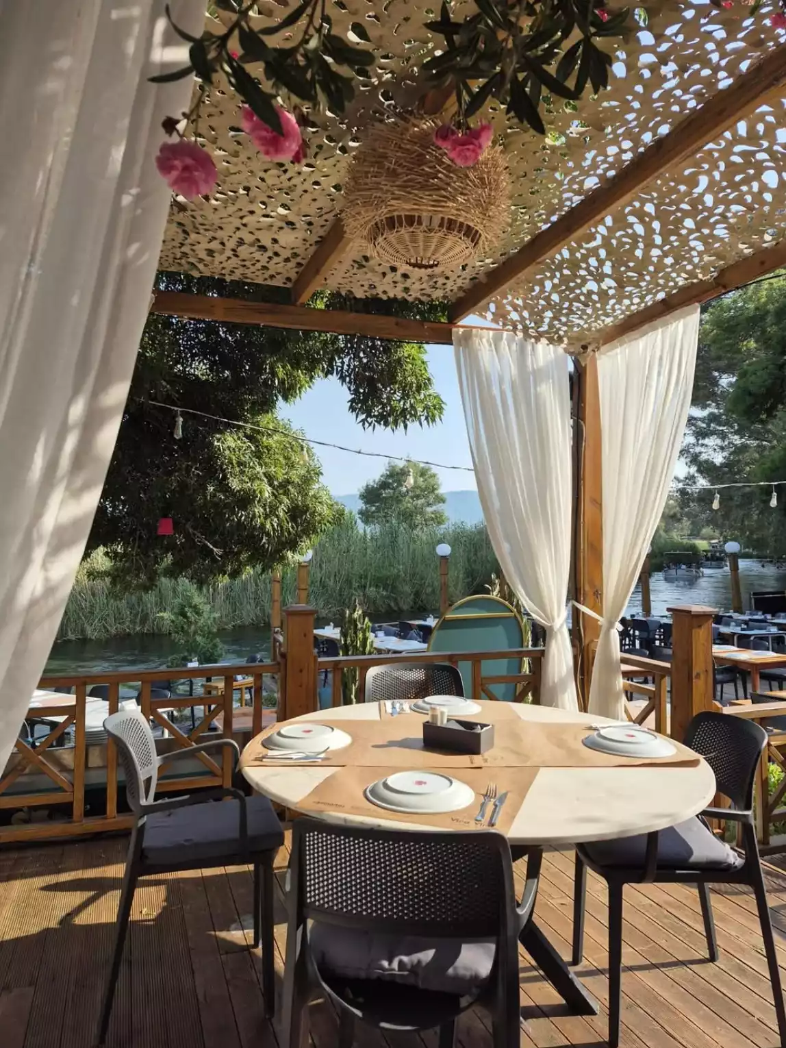 Akyaka'da, üzeri pembe çiçekler ve hasır avizeyle süslü pergolanın altında, su kenarında yeşillikler içindeki bir restoranın ahşap iskelesinde kurulmuş, hazır tabaklarla donatılmış yuvarlak bir masa ve sandalyeler davetkar bir atmosfer sunuyor.