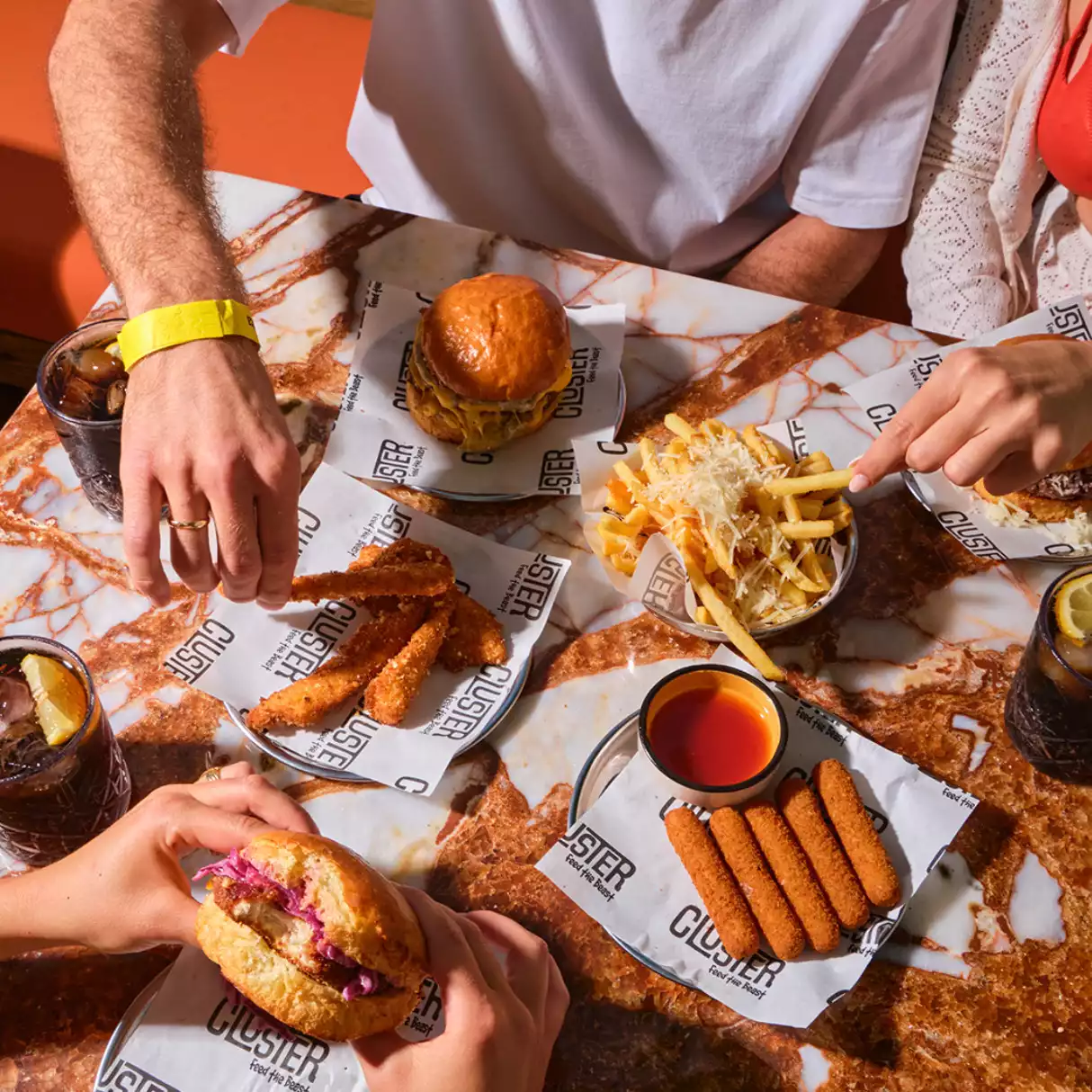 Mermer desenli bir masada toplanmış insanların Akyaka'da burger, peynirli patates kızartması, kızarmış tavuk ve çubuk peynir gibi çeşitli fast food lezzetlerini keyifle paylaştığı, bol atıştırmalıklarla dolu bir yemek masası görünümü.