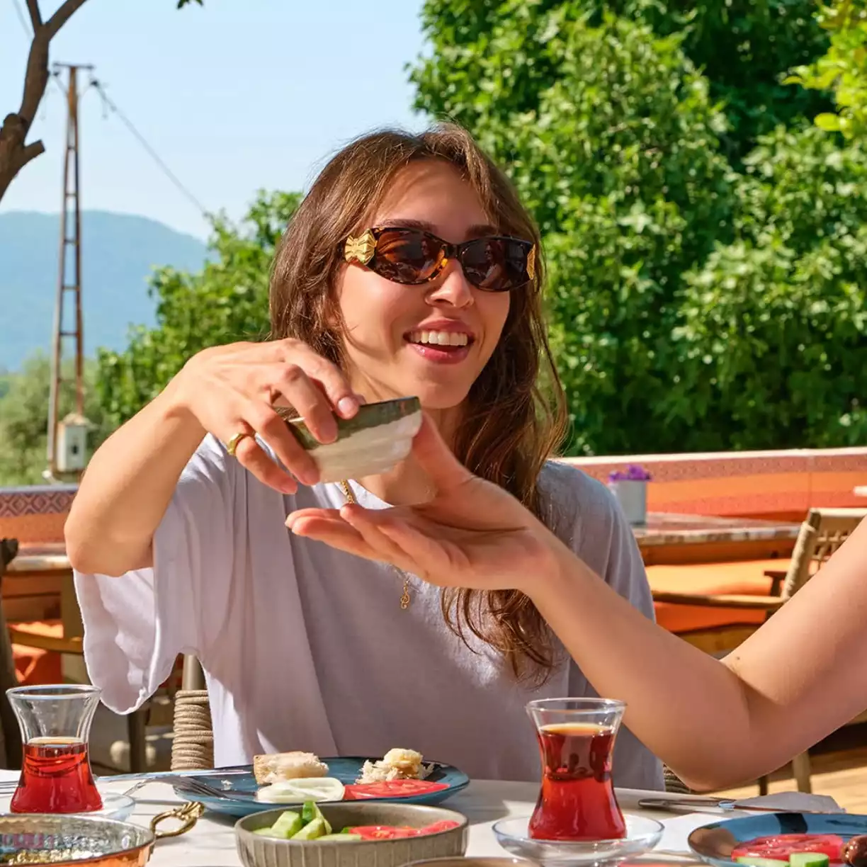 Akyaka'nın eşsiz doğal güzellikleri içinde, güneşli bir sabahın huzurlu atmosferinde, dostlarıyla geleneksel Türk çayı ve yöresel lezzetlerle dolu zengin bir kahvaltı sofrasında oturan, gözlüklü gülümseyen bir kadın, küçük bir kaseyi uzatırken keyifli anlar yaşıyor.
