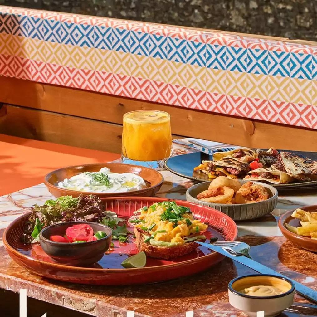 Akyaka'da mermer bir masada, üzerinde frenk soğanı serpilmiş çırpılmış yumurtalı ekmek, taze yeşil salata, yoğurtlu meze, bol iç harçlı dürüm benzeri yiyecekler, çıtır halka kızartmalar ve ferahlatıcı portakal suyunun bulunduğu zengin bir brunch veya kahvaltı sofrası, arka planda canlı renklerde desenli bir bank ile güneşli bir ortamda gösteriliyor.