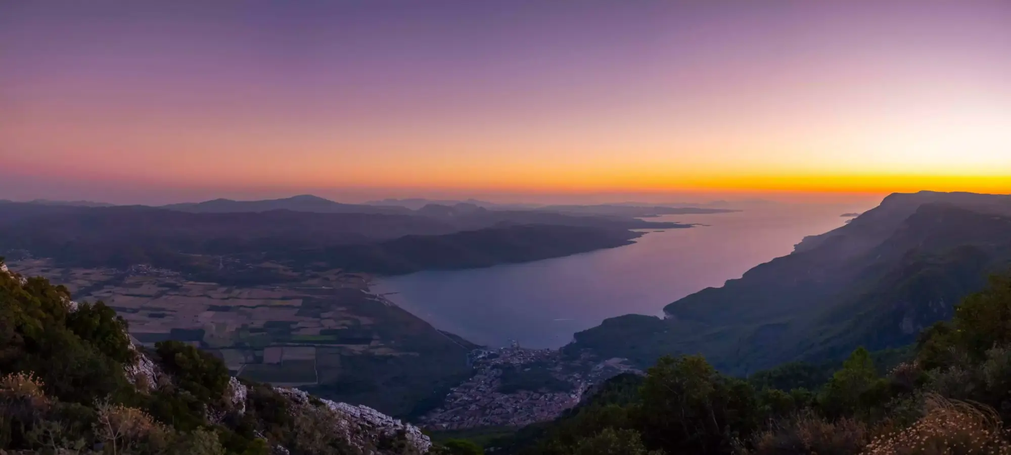 Akyaka koyu ve çevresindeki yerleşim yerlerini tepeden gösteren bu panoramik gün batımı fotoğrafında, mor ve turuncu tonlardaki gökyüzü, dingin deniz, kıyı şeridindeki ışıklı binalar ve yemyeşil tepelik araziler büyüleyici bir uyum sergiliyor.