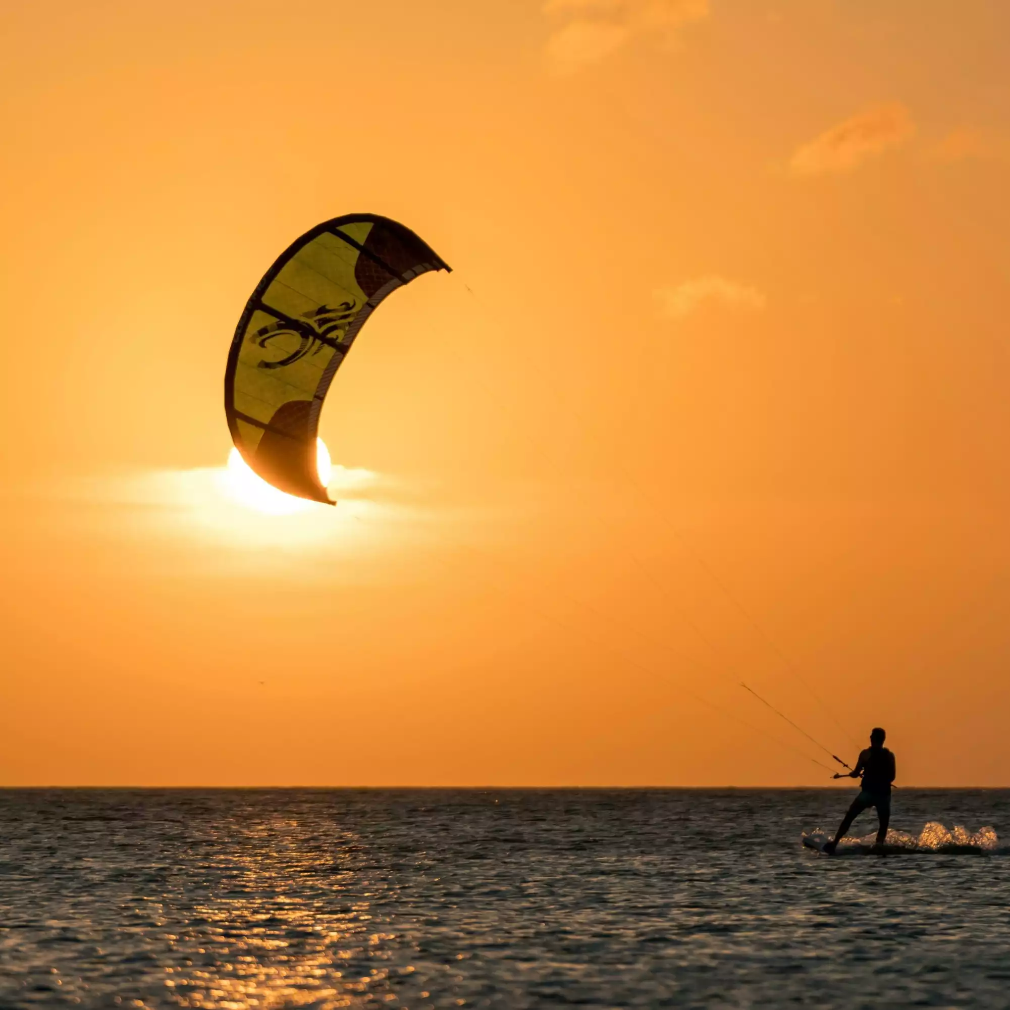 Akyaka'nın sakin sularında batan güneşin sıcak turuncu ışıkları altında, sarı-yeşil bir uçurtmayla denizde kitesurf yapan bir sporcu, ışıl ışıl parlayan denizin üzerinde enerjik bir şekilde ilerleyerek gün batımının eşsiz güzelliğiyle bütünleşiyor.