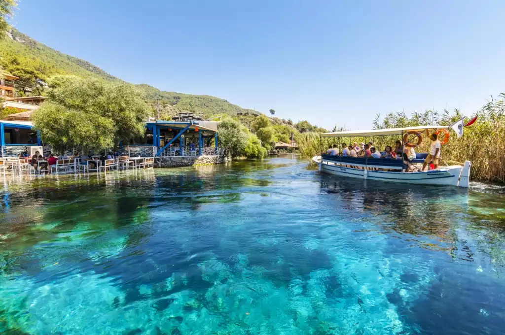 Akyaka'nın berrak turkuaz sularıyla ünlü Azmak Nehri'nde, güneşli bir günde tekne turu yapan kalabalık turist grubu ve sol kıyıda yemyeşil ağaçlar altında nehrin üzerine kurulmuş, keyifli anlar geçiren misafirlerle dolu bir restoran net bir şekilde görülmektedir.