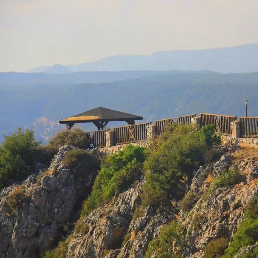 Akyaka'nın kayalık bir yamacında yer alan, ahşap korkuluklarla çevrili bir seyir terası ve çatılı bir kameriye, uzaklardaki dağlar ve vadinin nefes kesen panoramik manzarası eşliğinde görülüyor.