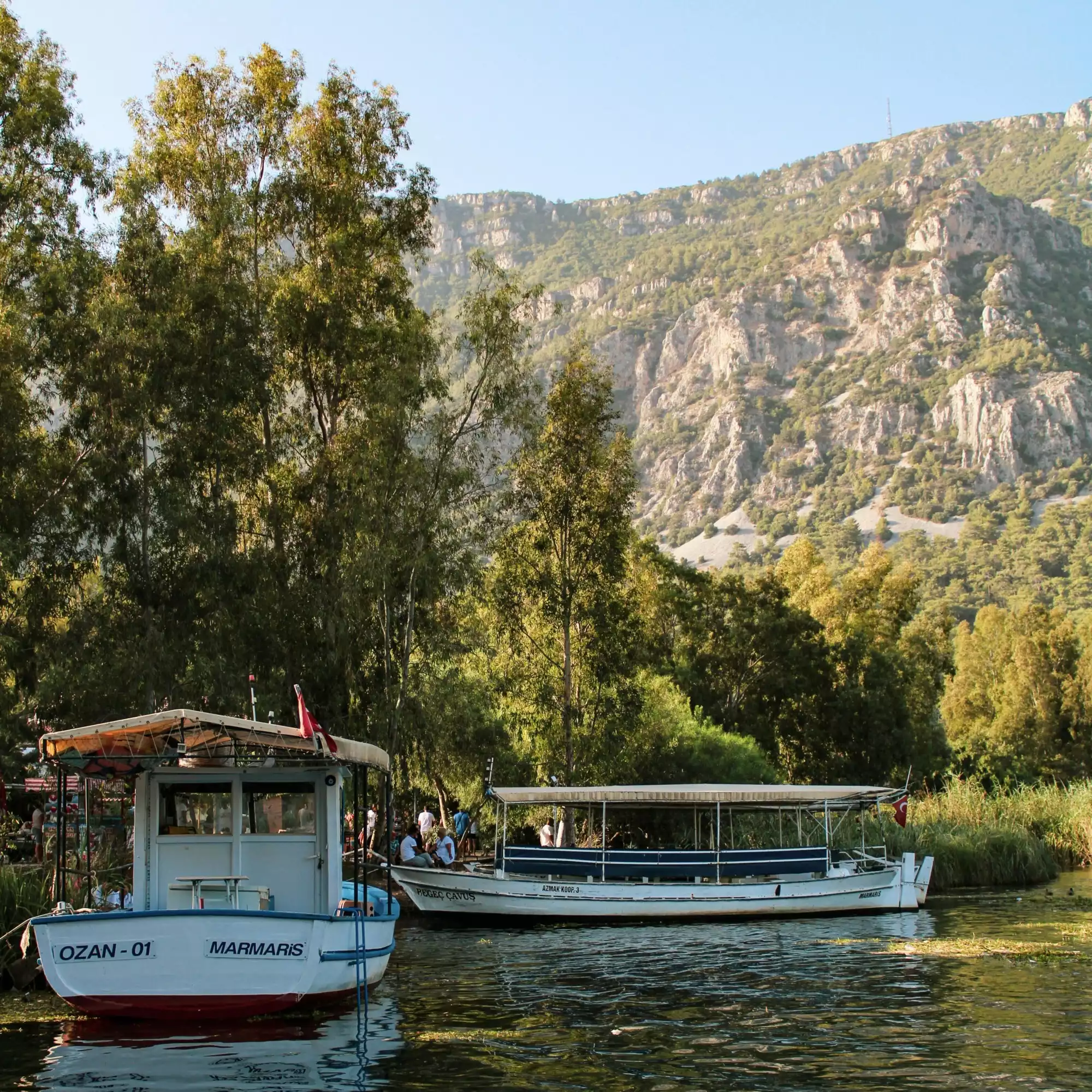 Akyaka'nın meşhur Azmak Nehri'nde, yemyeşil ağaçlar ve heybetli kayalık dağlarla çevrili sakin sularda, üzerinde Ozan-01 Marmaris yazan küçük bir tekne ve içinde yolcularıyla bekleyen uzun bir tur teknesi yer alıyor.