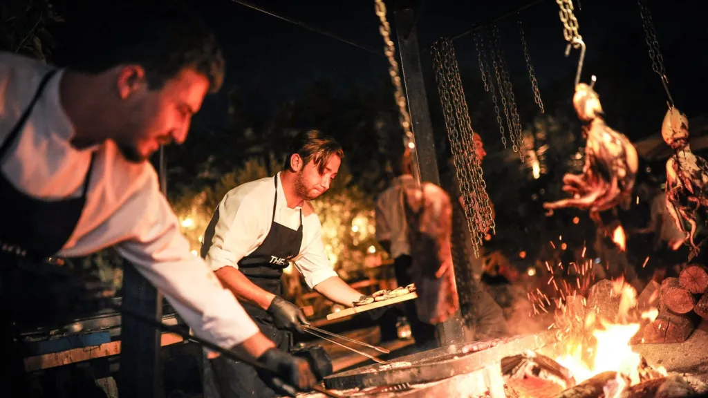 Gece açık havada, The Stay Warehouse'un şefleri, közlerin ve alevlerin parladığı büyük bir Arjantin usulü ızgaranın başında, zincirlerden asılı etleri yavaşça pişirirken, diğer yiyecekleri de özenle hazırlıyorlar.
